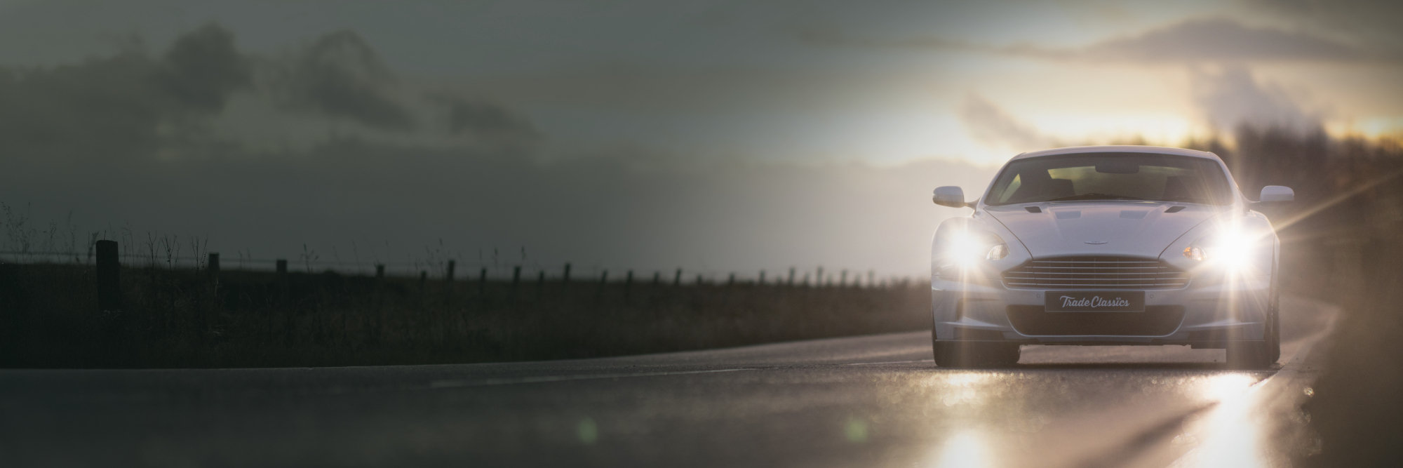 An Aston Martin classic car driving on a country road with headlights on at dusk